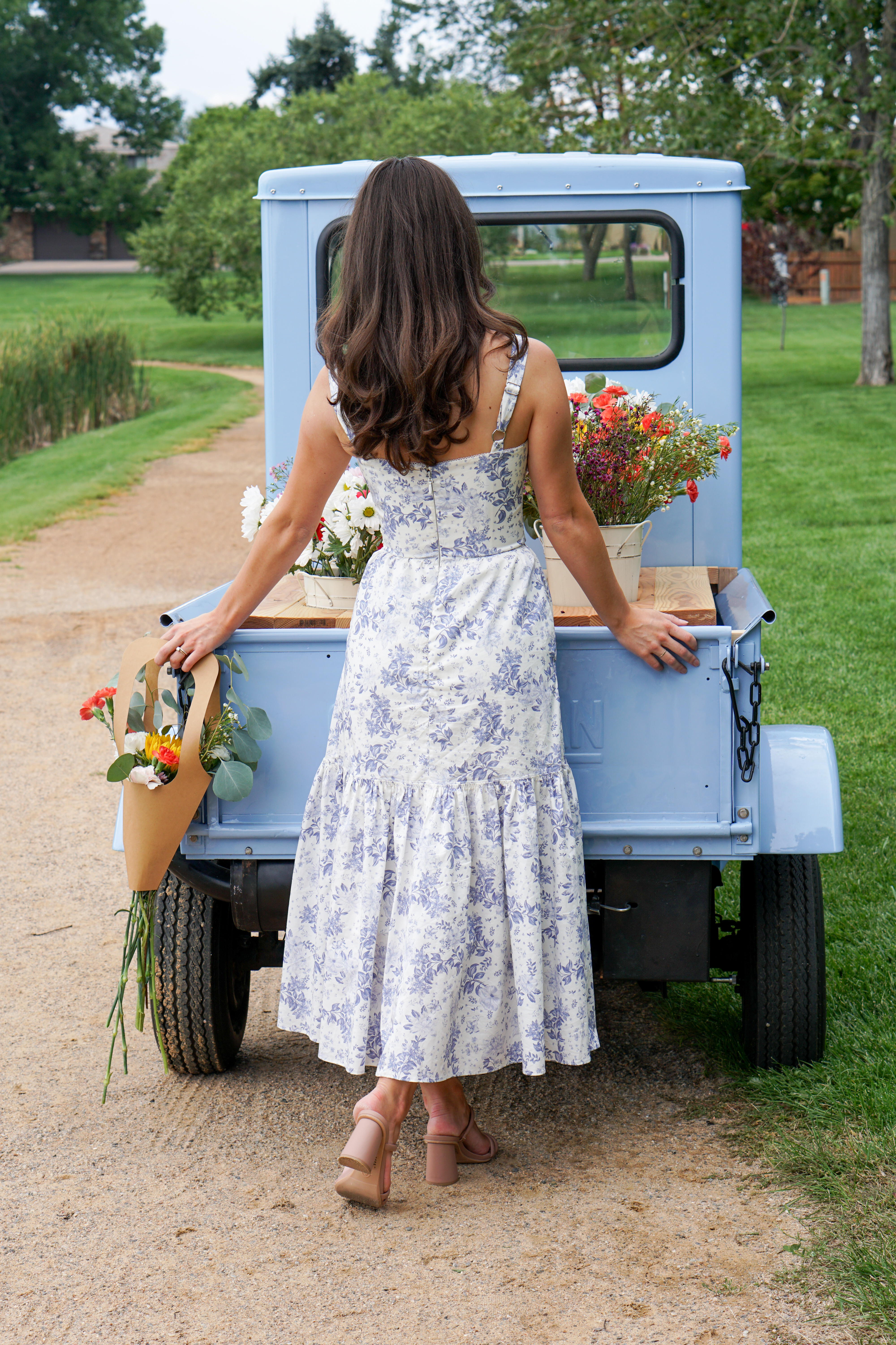 A woman in a floral dress stands beside a light blue vintage truck, holding a bouquet of flowers and admiring the view in a grassy outdoor setting.