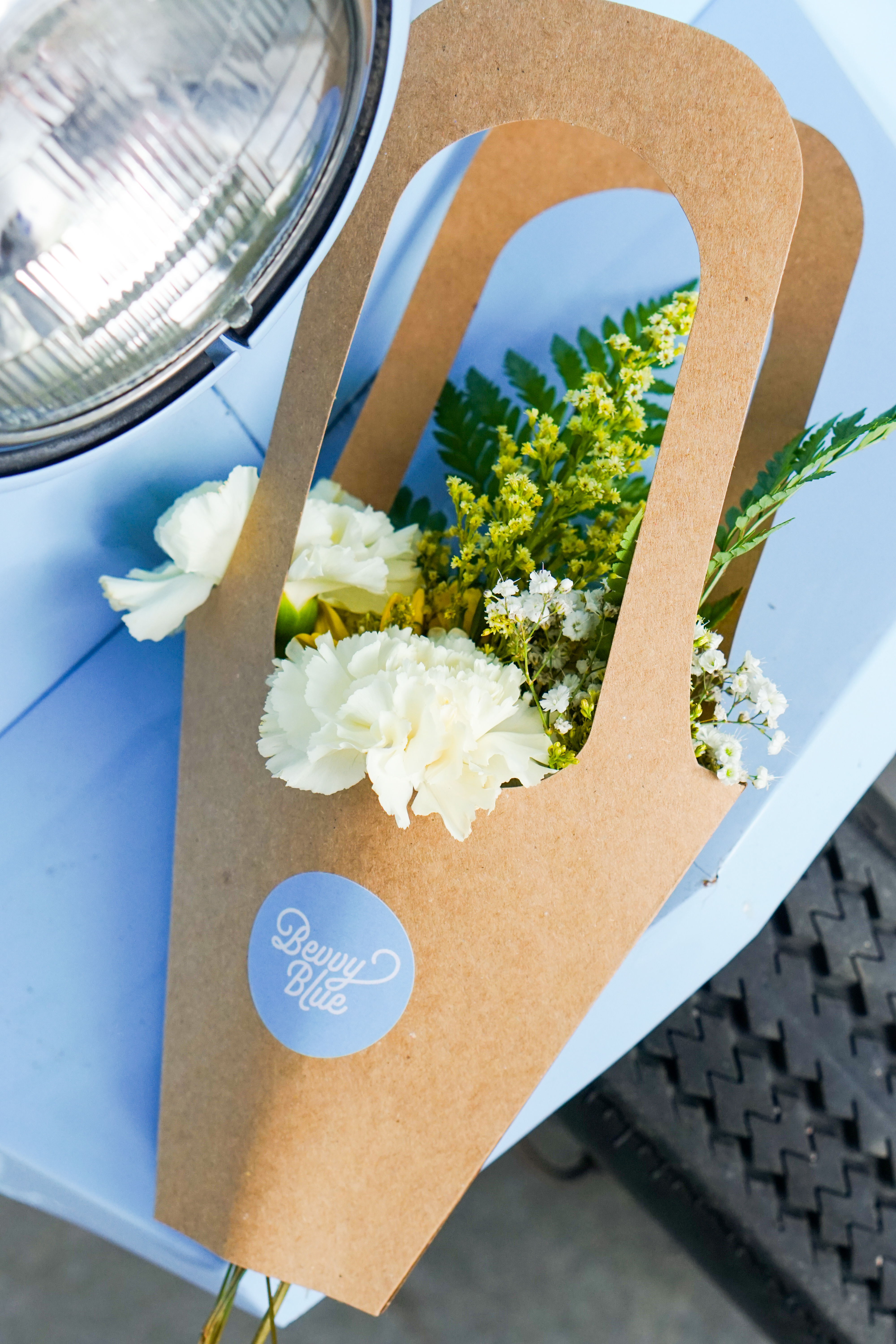 A bouquet of mixed flowers including white carnations and greenery, presented in a brown paper wrap with a blue logo label on a light blue surface.