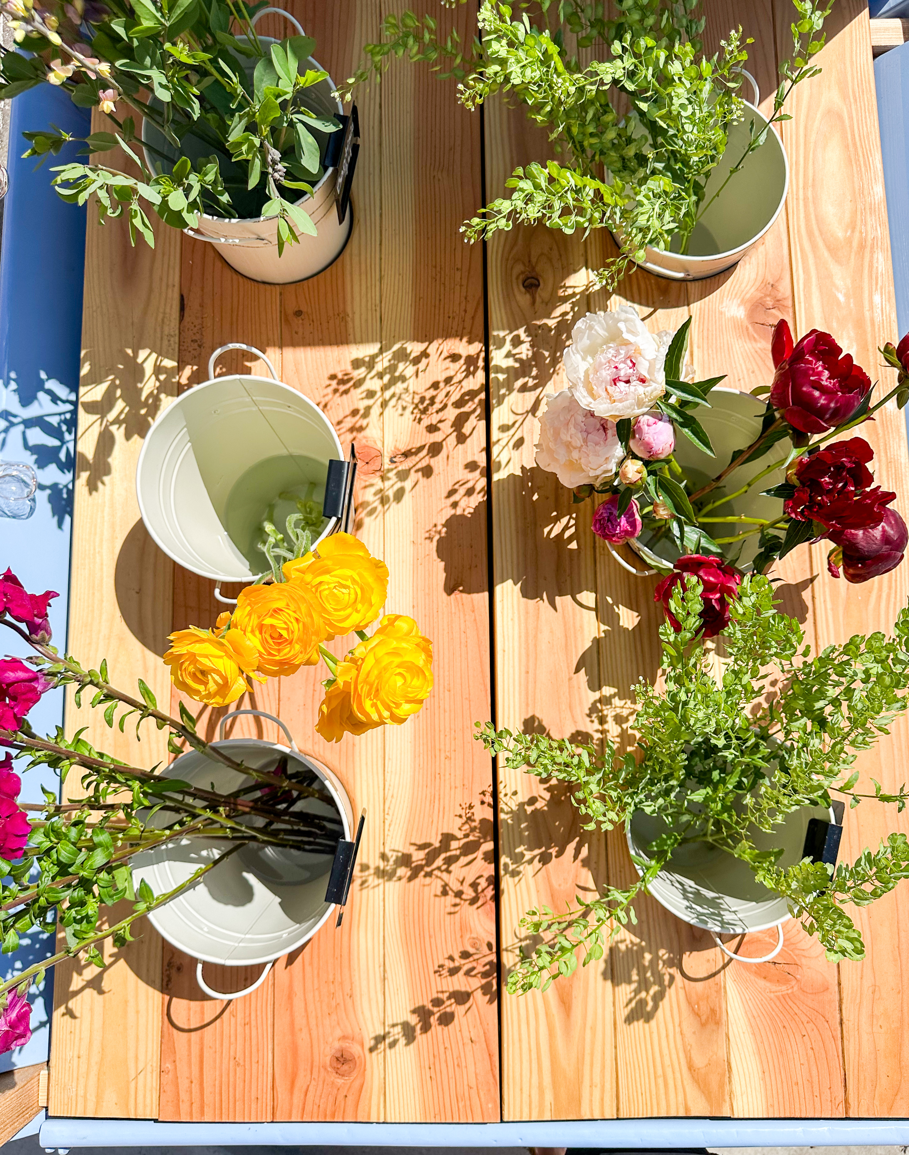 The truck bed of a cushman truckster filled with several small buckets filled with colorful flowers, including yellow ranunculus, white peonies, and red flowers, surrounded by greenery, illuminated by sunlight.